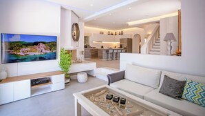 A modern living room with a flat-screen TV, a coffee table with decorative items, a sofa with cushions, and a kitchen area in the background. - VILLA PANAMERA,  Infinite view, Private heaven (Mykonos)