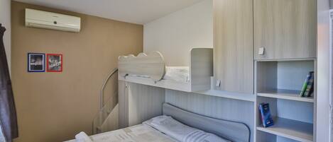 A bedroom with a bed, a wardrobe, a shelf with books, and an air conditioning unit.