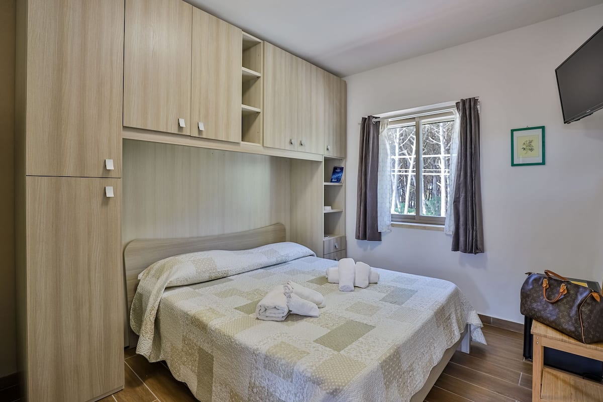 Ein ordentlich eingerichtetes Schlafzimmer mit Bett, Kleiderschrank, einem Fenster mit Vorhängen und einem Fernseher an der Wand.