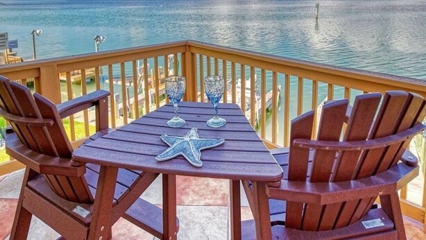 Eine Holzterrasse mit Tisch und Stühlen, die auf ein ruhiges Meer mit einer entfernten Küste blickt.