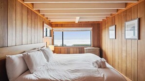 Une chambre avec un mur lambrissé en bois, un grand lit avec une literie blanche, une fenêtre donnant sur un beau paysage et des tableaux encadrés accrochés au mur.
