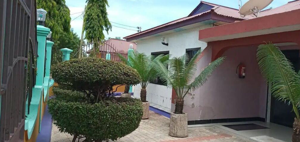 Un quartier résidentiel avec une maison dotée d’une antenne parabolique, d’un toit rouge et d’un petit jardin agrémenté de plantes en pot.