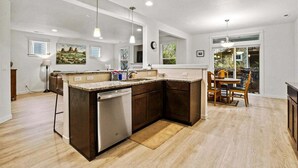 Una cocina moderna con una isla central, armarios de madera y lavavajillas. Hay luces colgantes, un comedor con mesa y sillas, y vistas al exterior con vegetación.