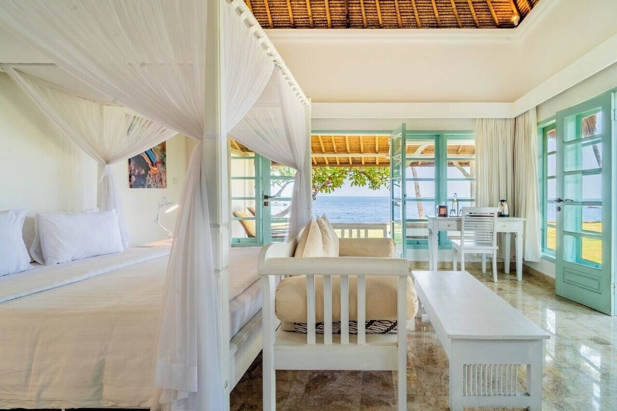 Quarto com cama, um banquinho, uma escrivaninha e vista para o mar.