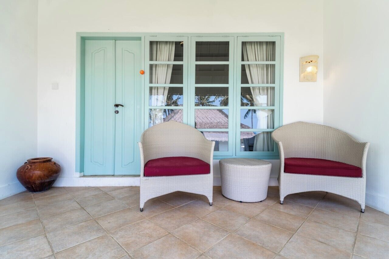 A room with two wicker chairs with red cushions, a small round table, a large window with curtains, and a light blue door.