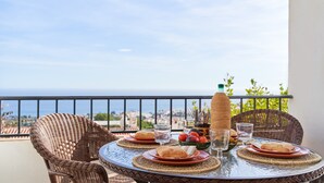 Une table ronde, avec des assiettes, des verres et une bouteille, est placée sur un balcon donnant sur la ville et la mer.