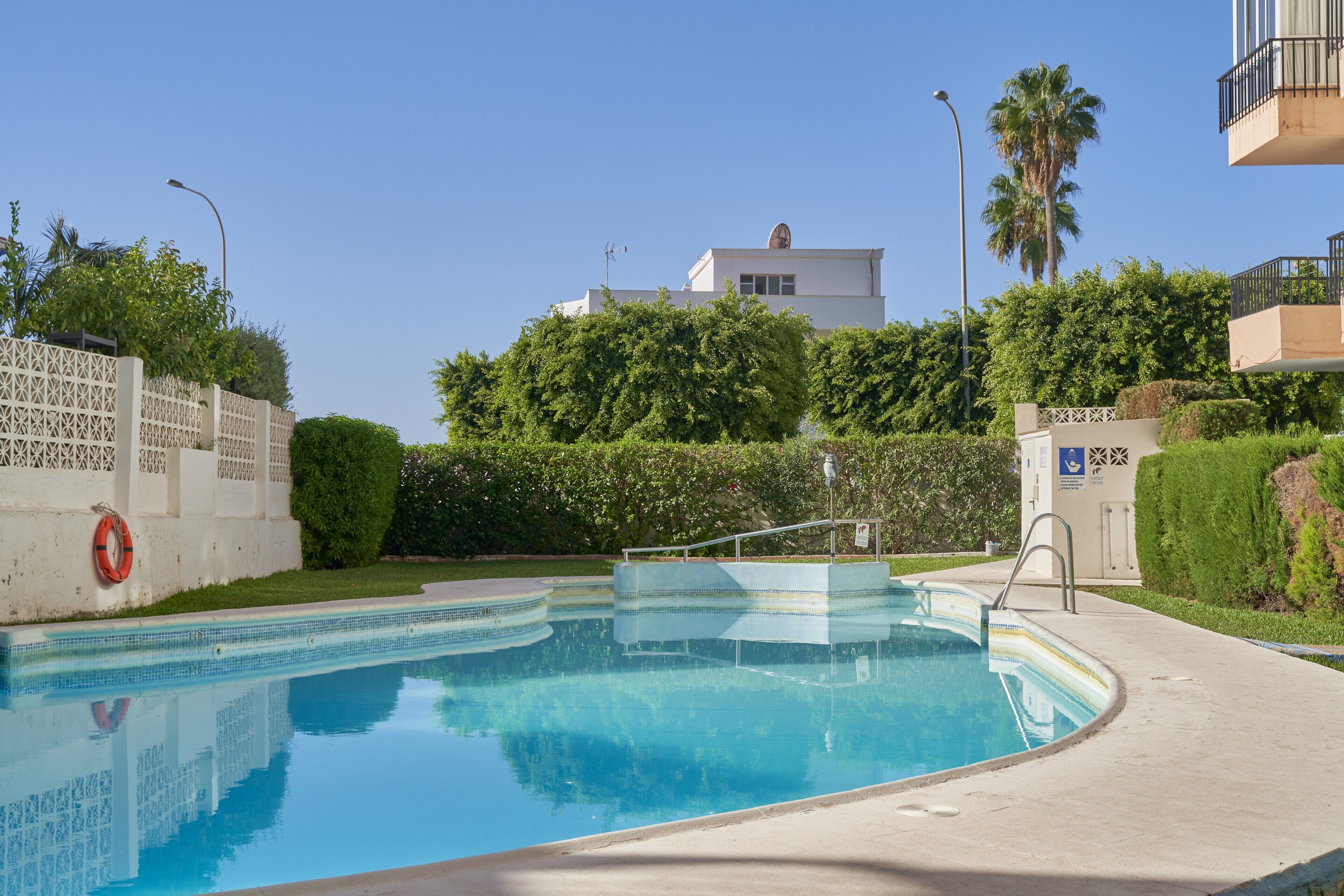 Une piscine à l’eau d’un bleu limpide, entourée d’une clôture blanche, avec un bâtiment visible au loin.