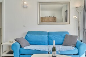 Un canapé bleu, avec un miroir au-dessus, une petite table sur laquelle se trouvent une bouteille et un verre, et une lampe sur la droite.