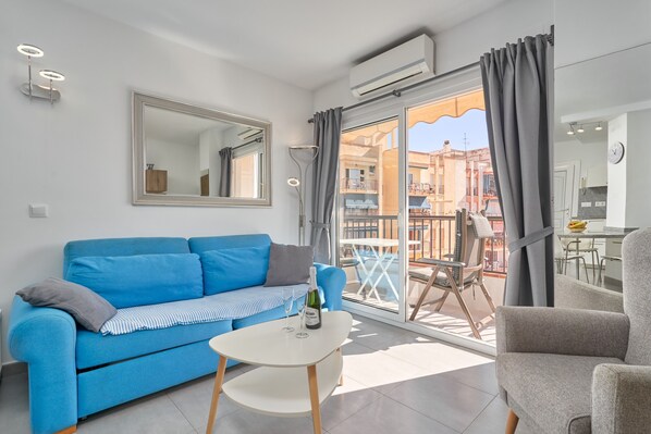 Un salon moderne avec un canapé bleu, une petite table blanche sur laquelle se trouvent une bouteille et des verres, un grand miroir et un balcon offrant une vue sur les immebles.