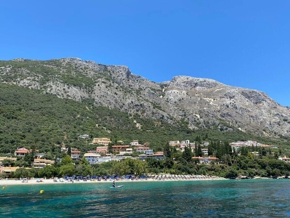 A coastal town with buildings along the beach, a clear blue sky, and a mountainous backdrop.