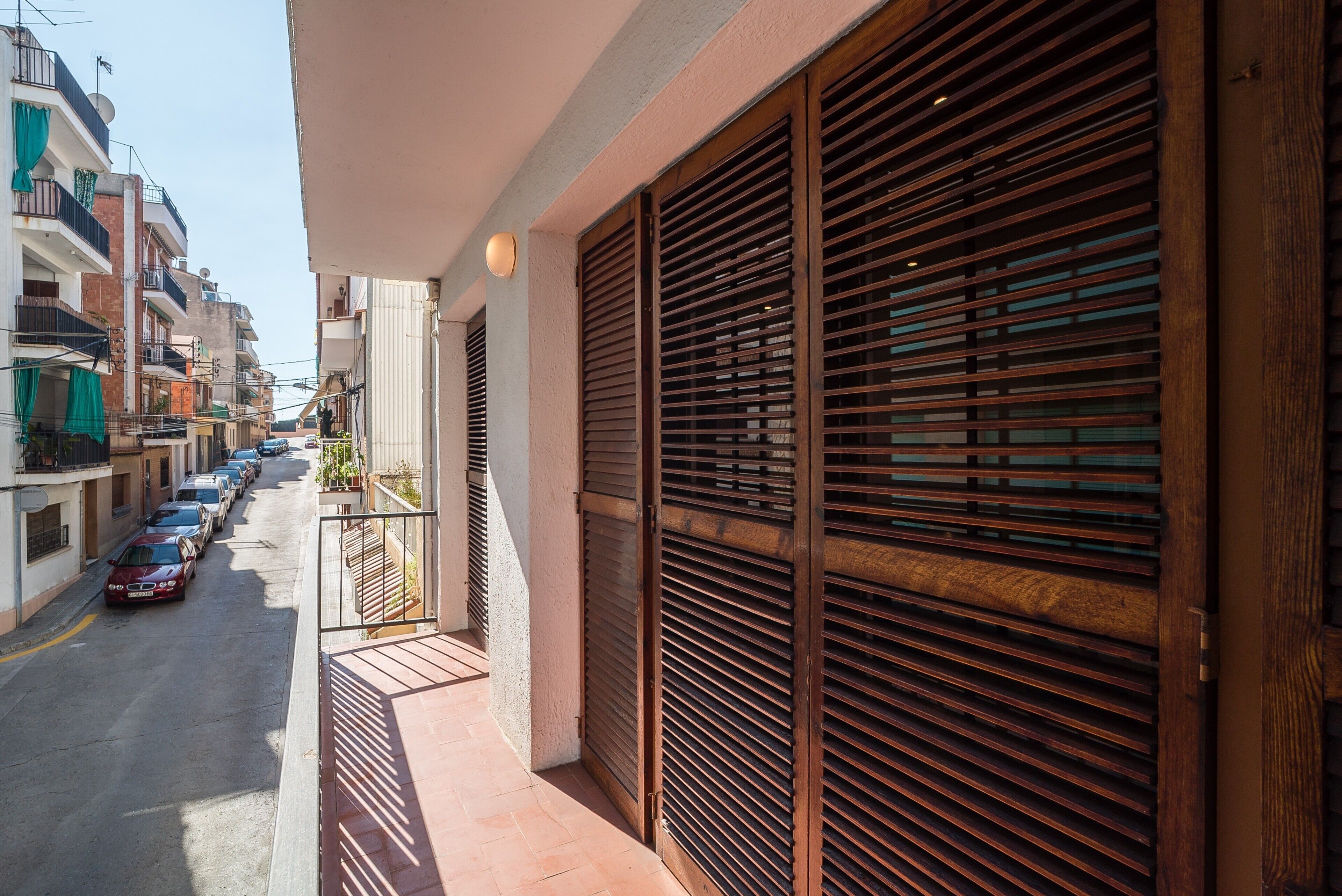 A narrow street with parked cars, a balcony with wooden shutters, and buildings with balconies and satellite dishes.