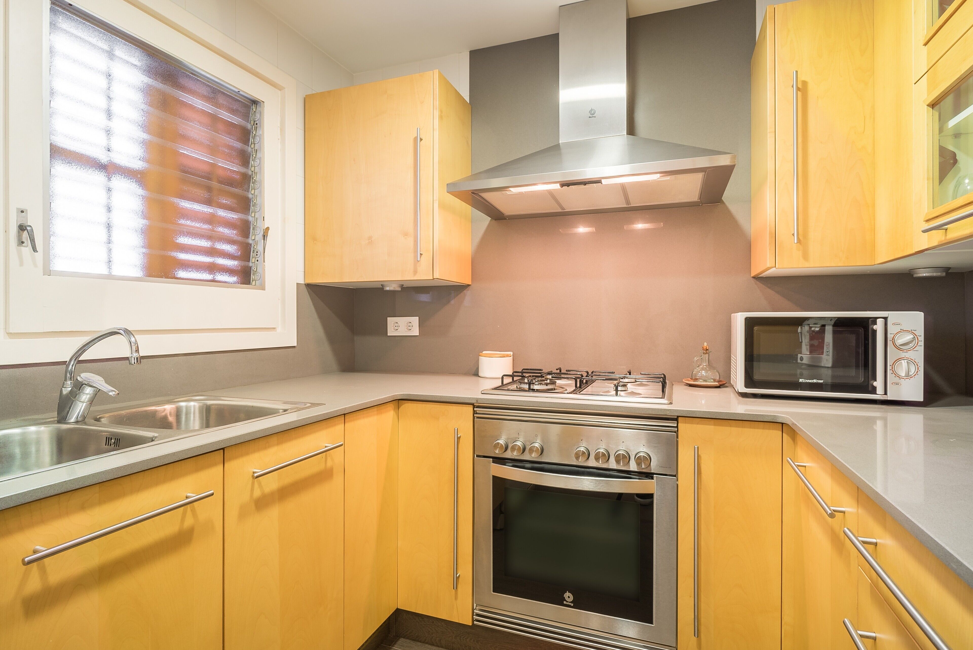 Een keuken met houten kastjes, een roestvrijstalen spoelbak, een gasfornuis, een oven en een magnetron.