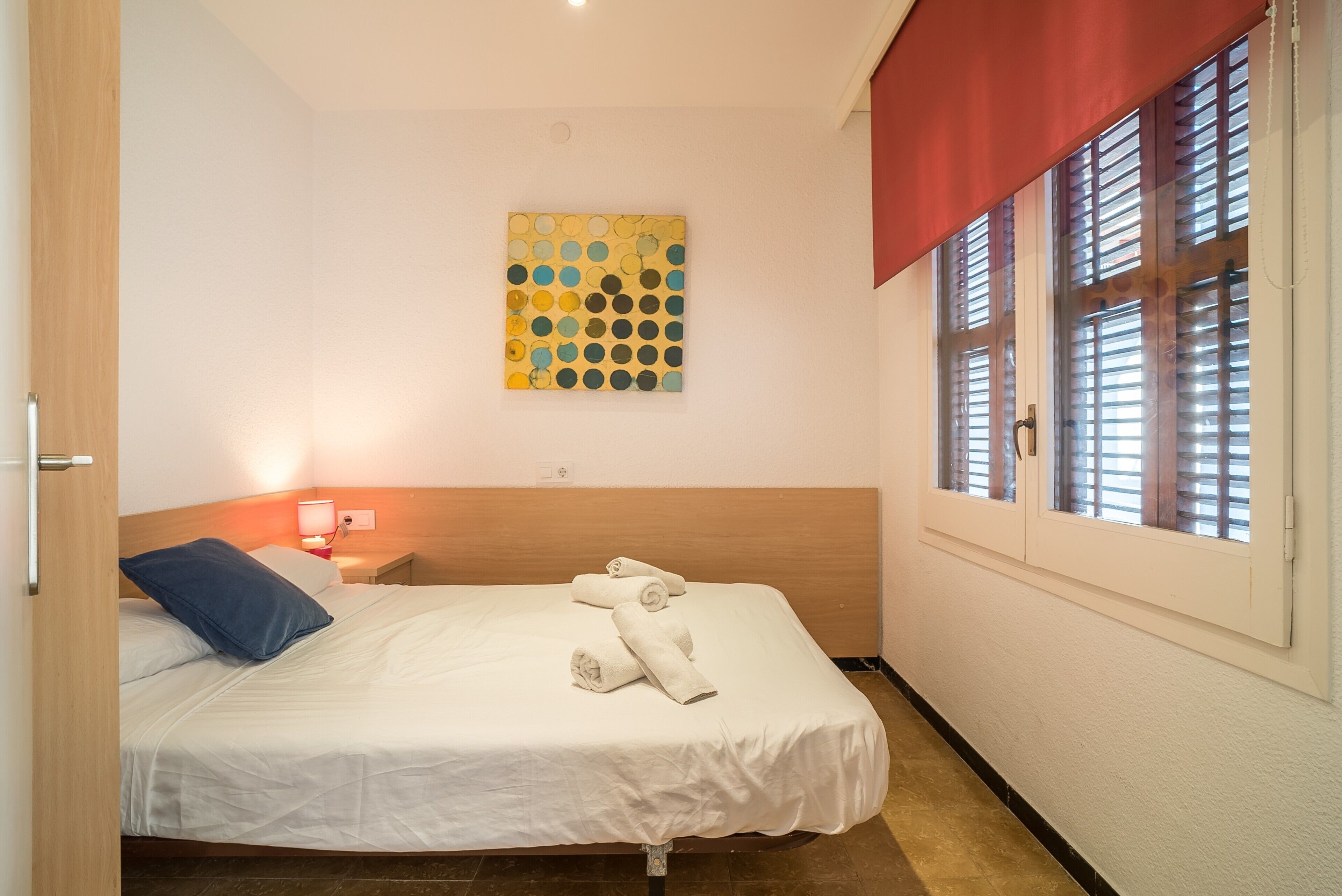 Een hotelkamer met een bed, een kunstwerk aan de muur, een raam met zonwering en een nachtkastje met een lamp.