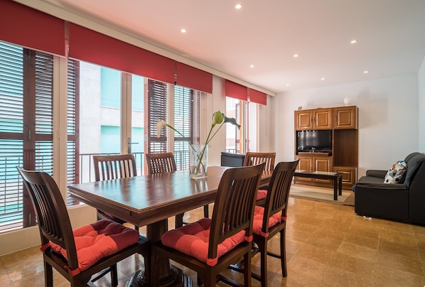 A dining area with a wooden table and chairs, a vase with flowers, and a kitchen area with cabinets and a TV.