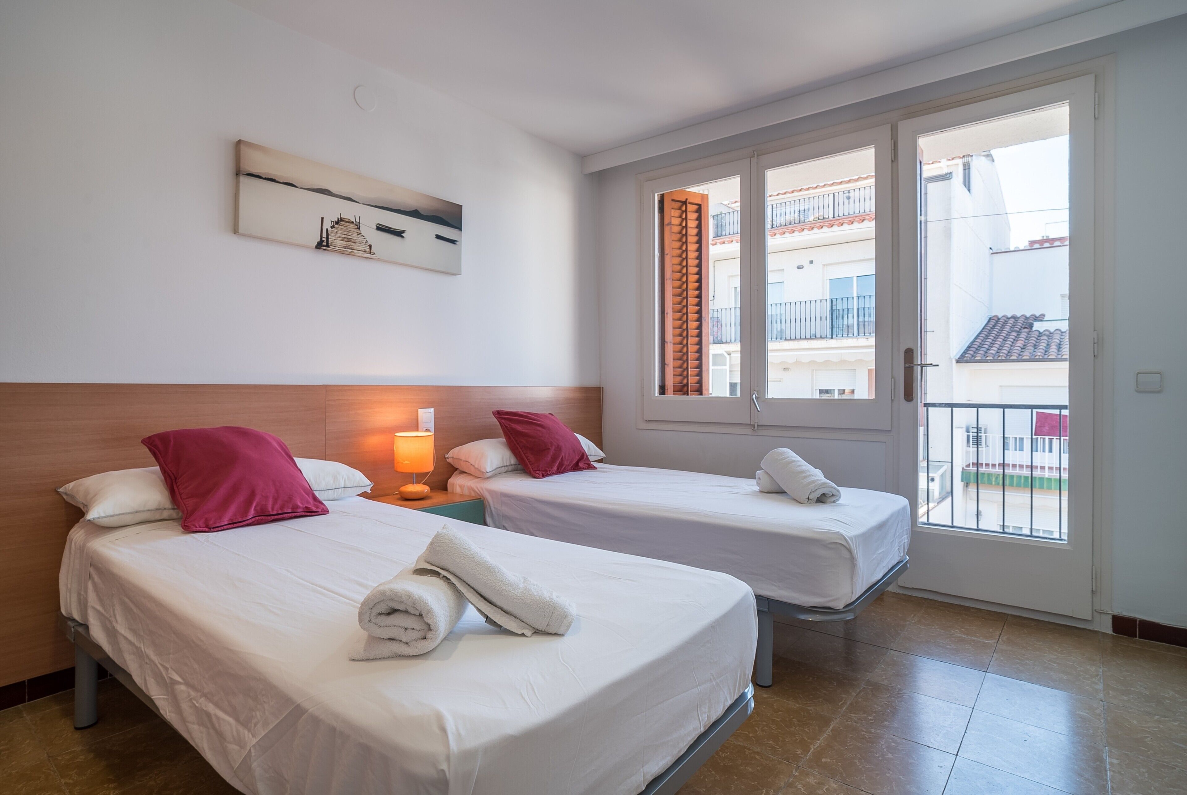 Habitación de hotel con dos camas, una mesita de noche con lámpara, un cuadro en la pared y un balcón con vistas a edificios.