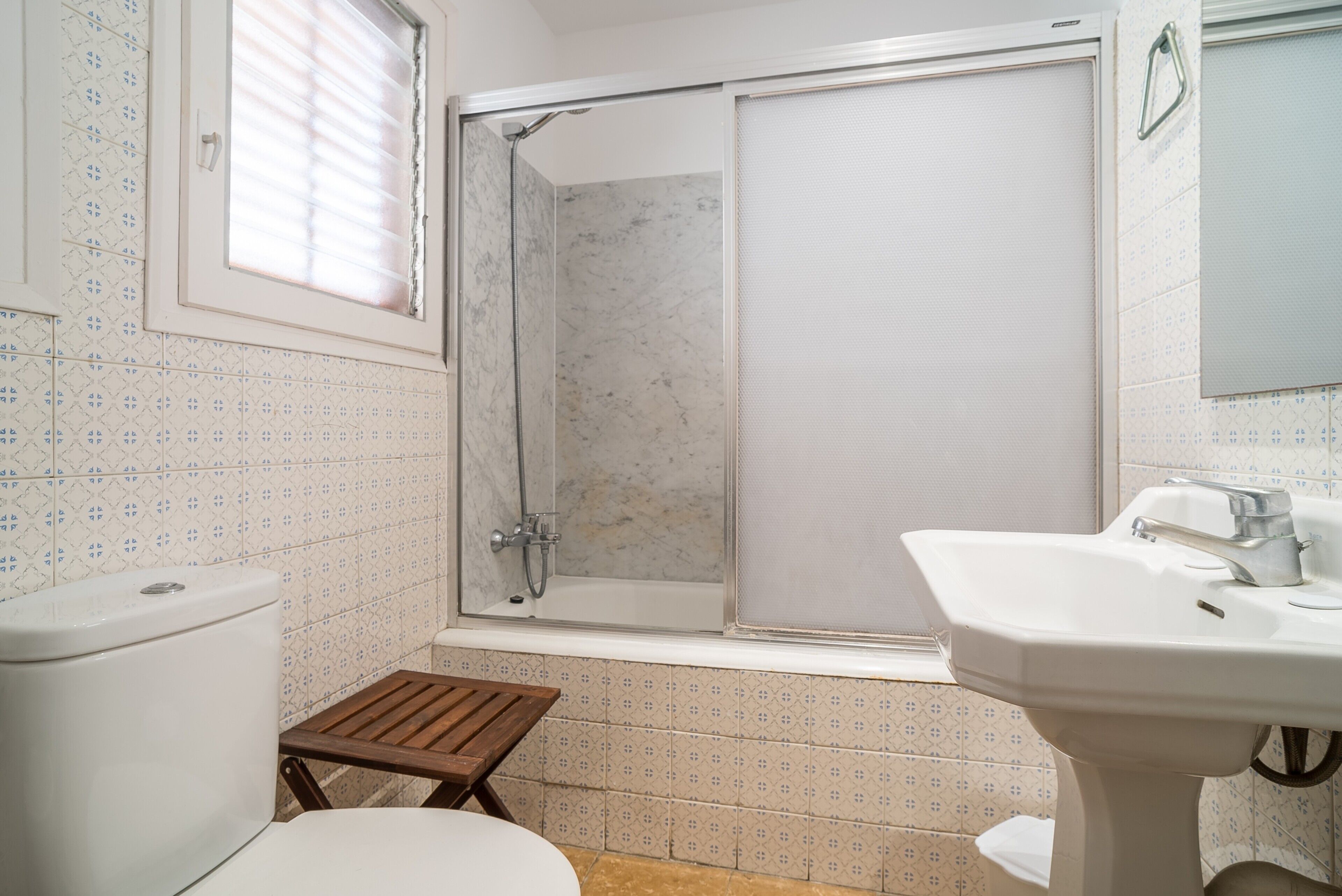 Un baño con inodoro, ducha con puerta de vidrio, lavamanos y un taburete de madera.