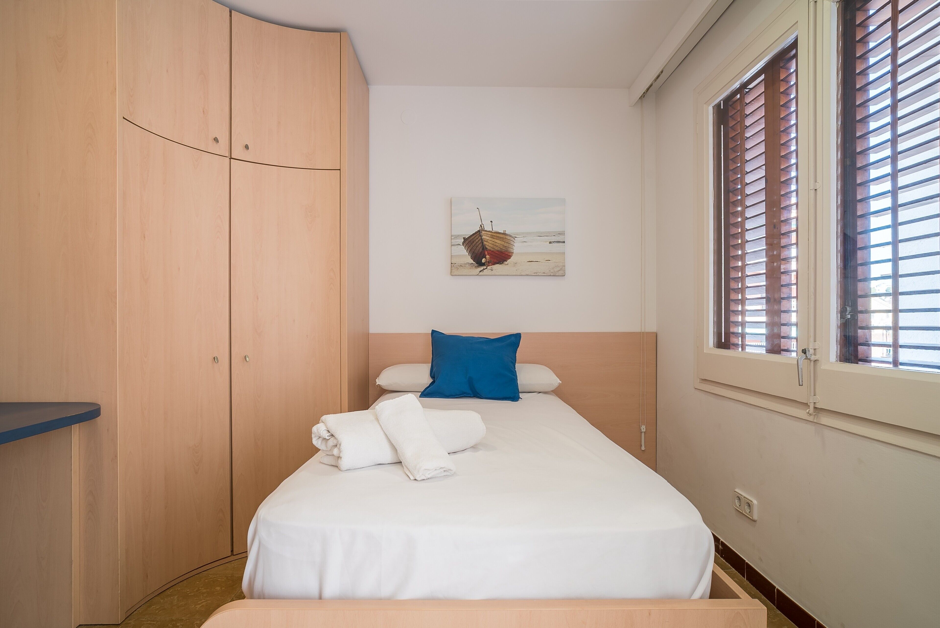 A bedroom with a bed, a wooden wardrobe, a window with blinds, and a painting on the wall.