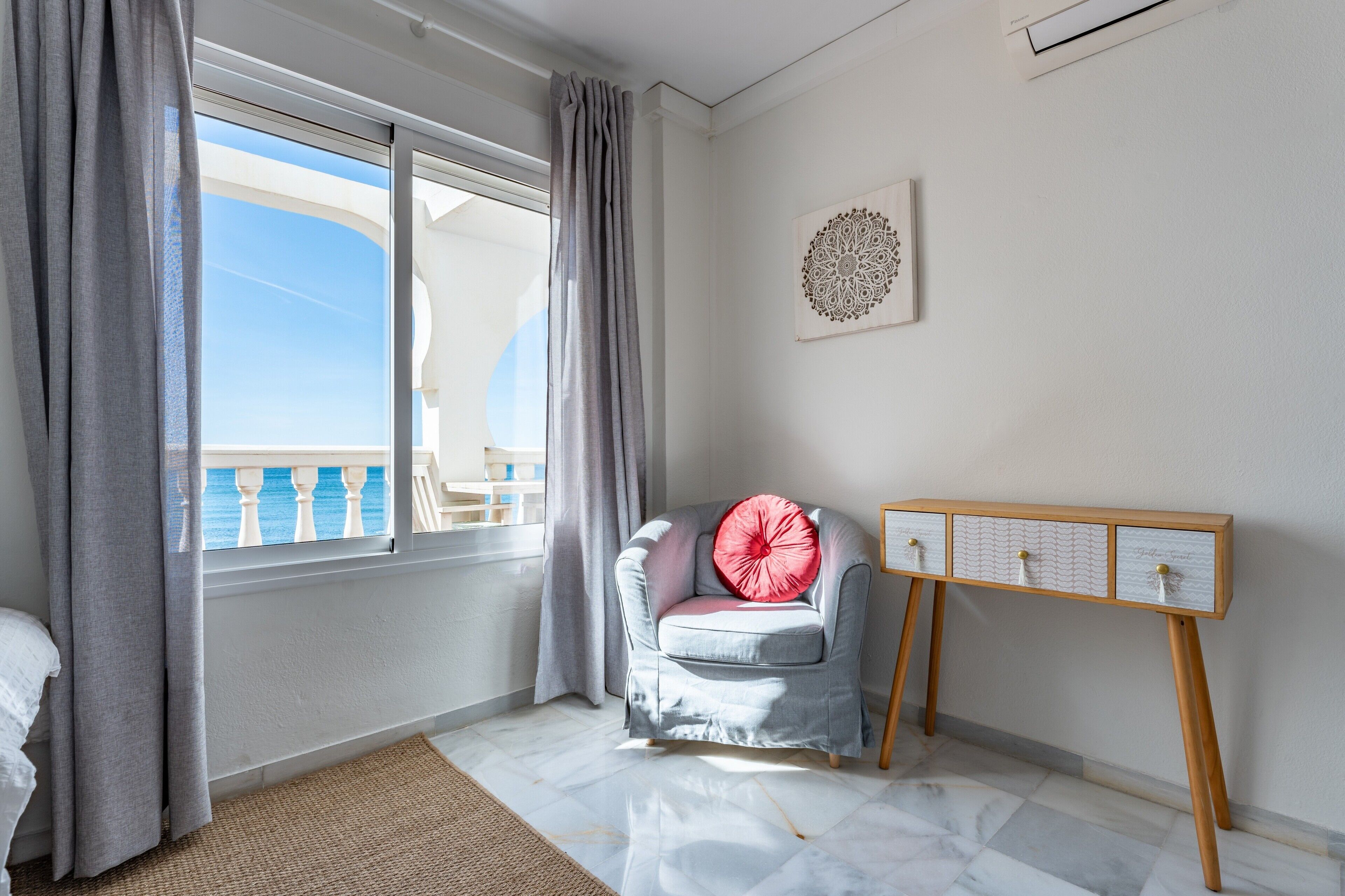 A room with a bed, a chair with a cushion, a console table, a decorative wall piece, and a view of the outside.