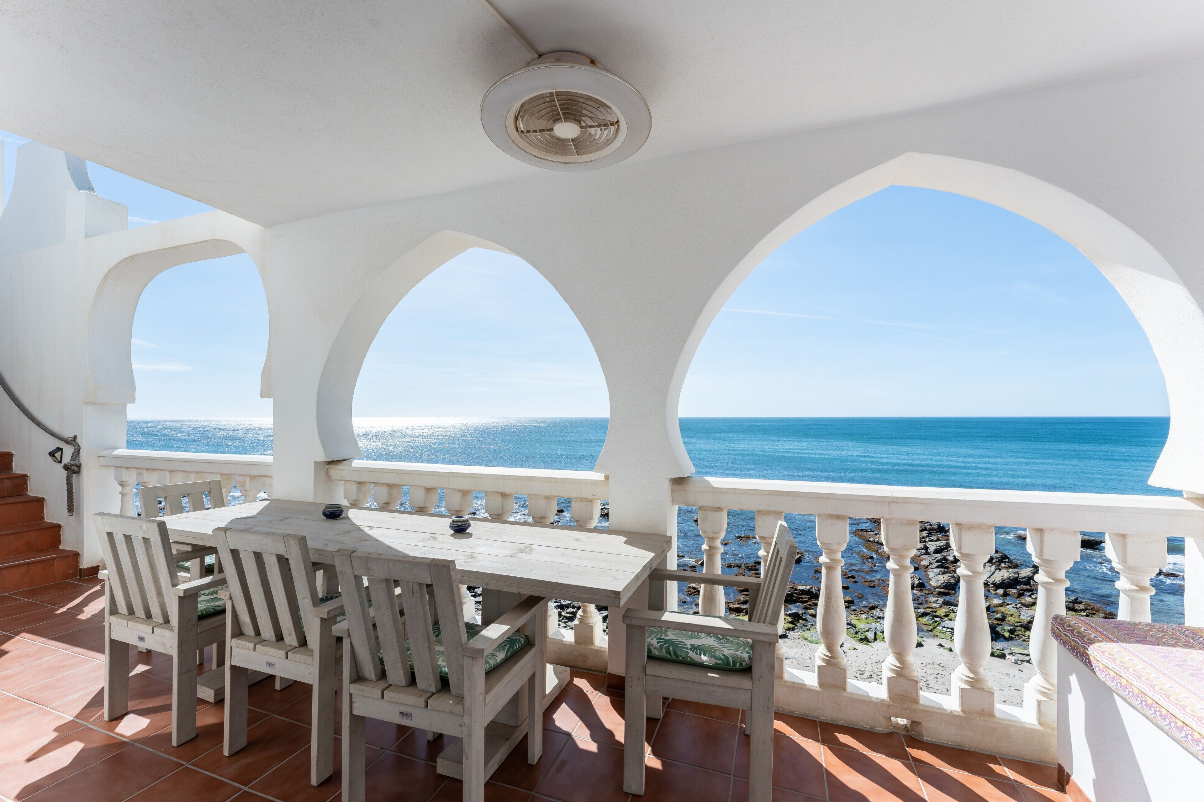 A terrace with a table and chairs, a ceiling fan, and a view of the sea.