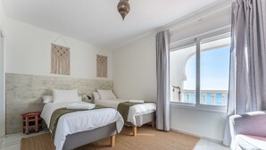 Habitación de hotel con dos camas, vista al mar, un balcón y un adorno de macramé colgando en la pared.