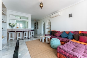 Ein modernes Wohnzimmer mit einer Couch, Hocker und einem kleinen Couchtisch. Im Hintergrund sind weiße Barhocker und eine Küchenzeile zu sehen.
