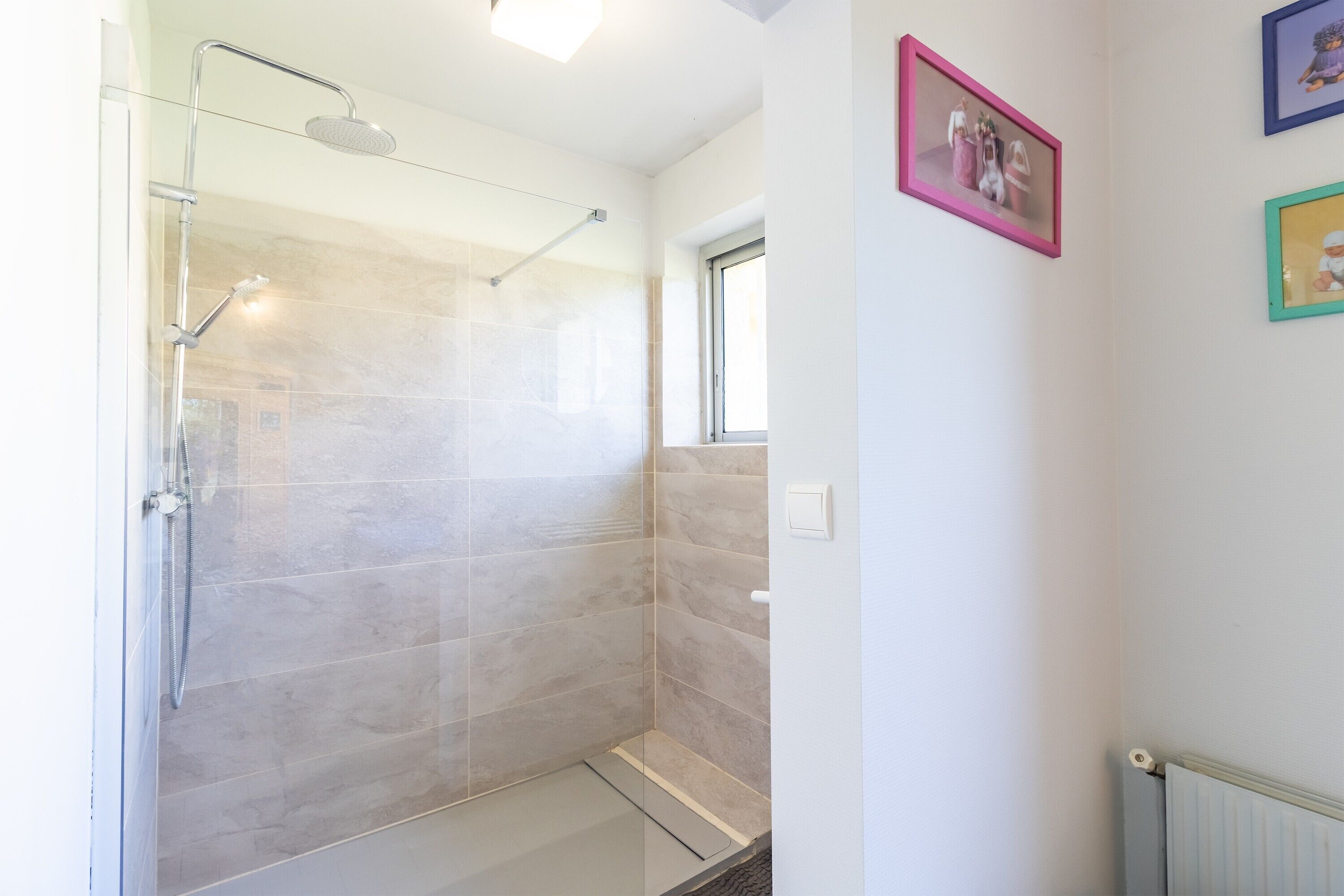 A glass-enclosed shower with a handheld showerhead, a window, and a radiator on the wall.