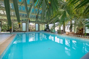 A clear indoor swimming pool with a glass ceiling, surrounded by palm trees and a view of the outdoors. - Spacious holiday home with indoor pool, jacuzzi and sauna in Cherveix-Cubas (Cherveix-Cubas)
