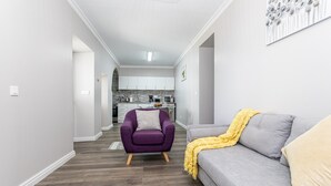 Un salon moderne avec un fauteuil violet, un canapé gris agrémenté d’un plaid jaune, et une cuisine en arrière-plan.