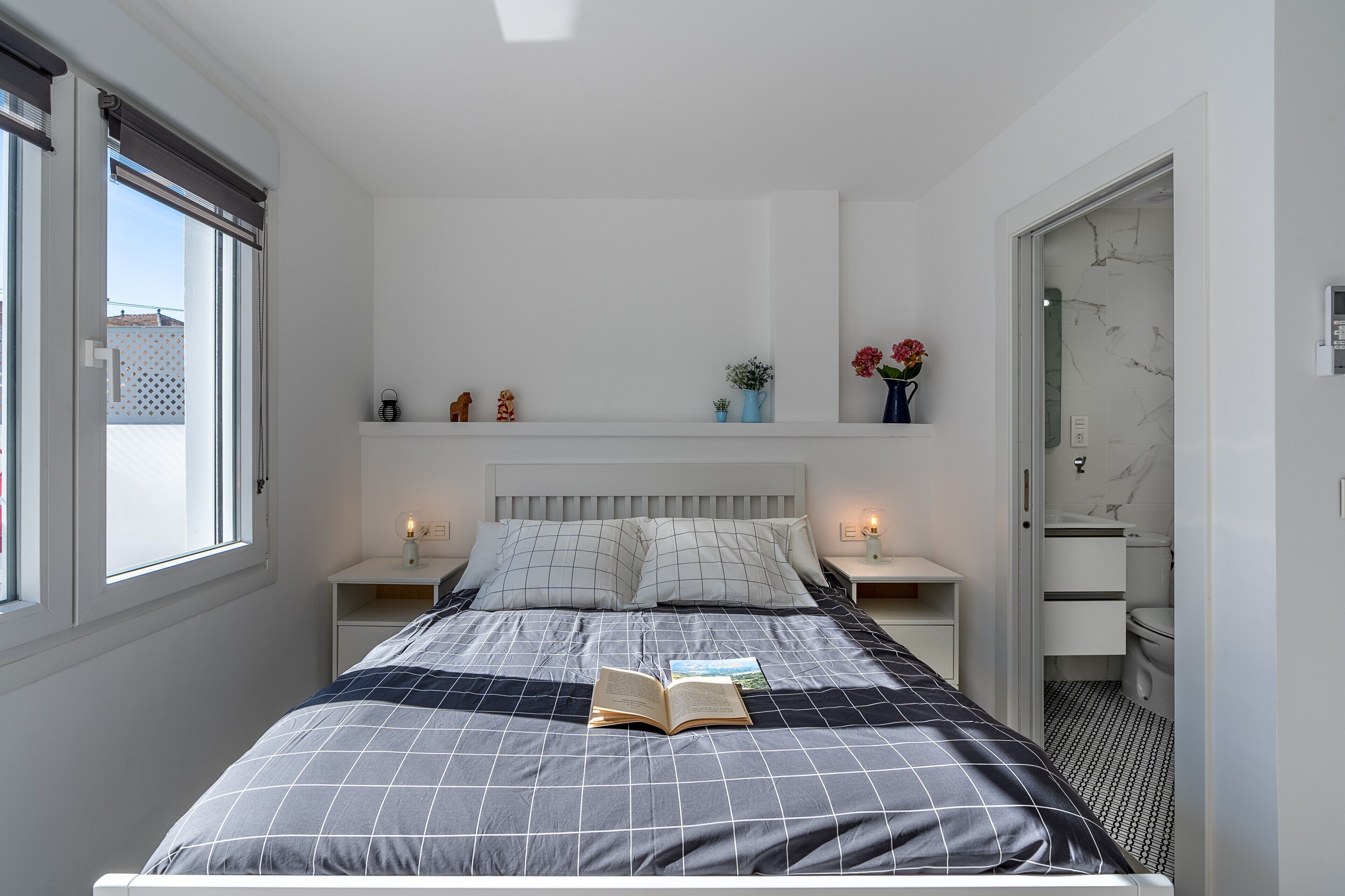 Een slaapkamer met een bed, een nachtkastje, een raam, een plank met decoratieve objecten en een deur naar de badkamer.