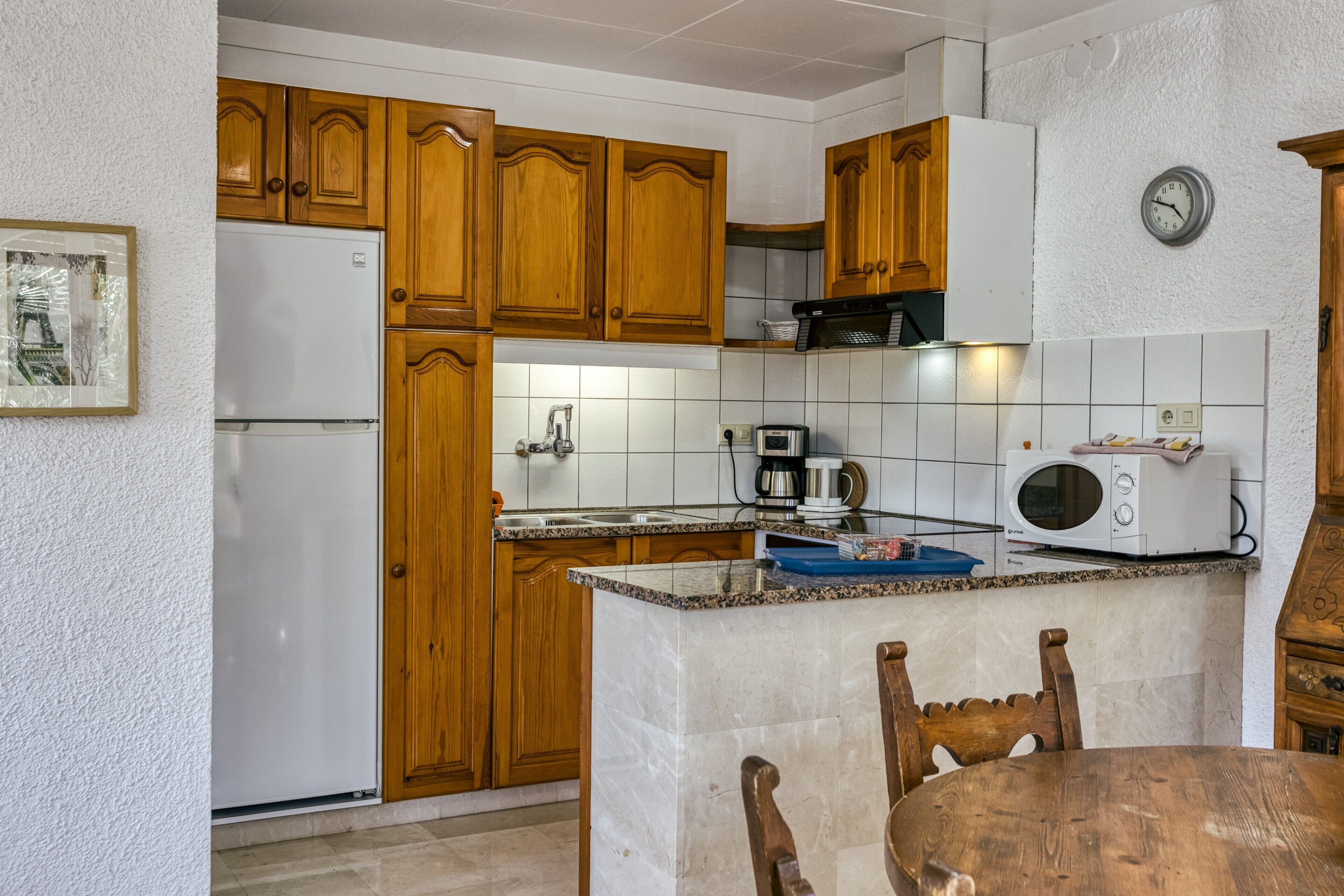 Une cuisine avec des meubles de rangement en bois, un réfrigérateur blanc, un four à micro-ondes et une horloge au mur.