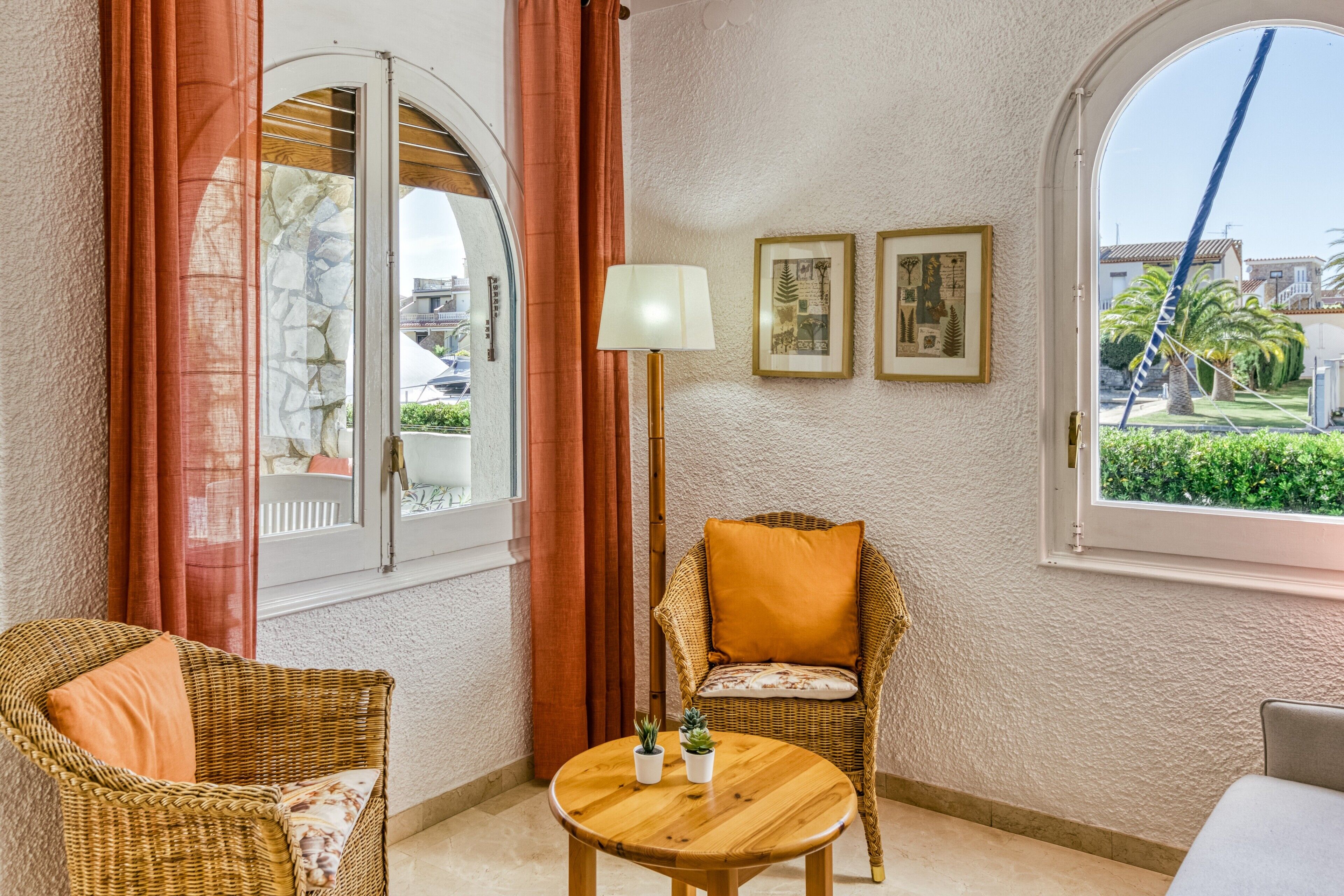 A room with a wicker chair, a round wooden table, a lamp, and framed pictures on the wall.
