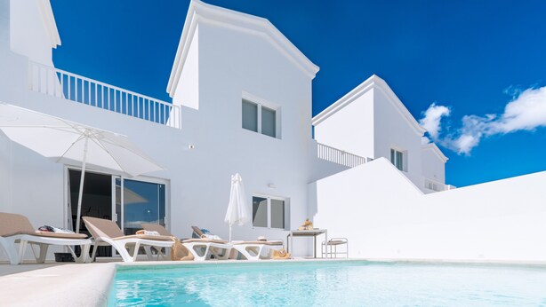 Une villa blanche avec une piscine, des chaises longues et une table ombragée par des parasols.
