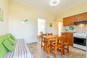 Une pièce comprenant une table à manger, des chaises, un canapé avec des coussins verts, un coin cuisine avec une cuisinière et des placards, ainsi qu’un mur recouvert de carreaux.