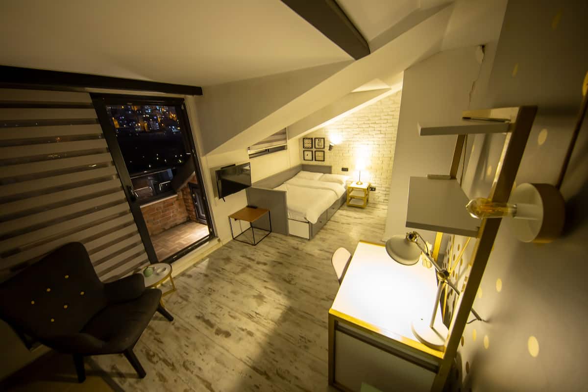 Un dormitorio con cama alta, escritorio y silla. Se ve una ventana con vista a la ciudad de noche.