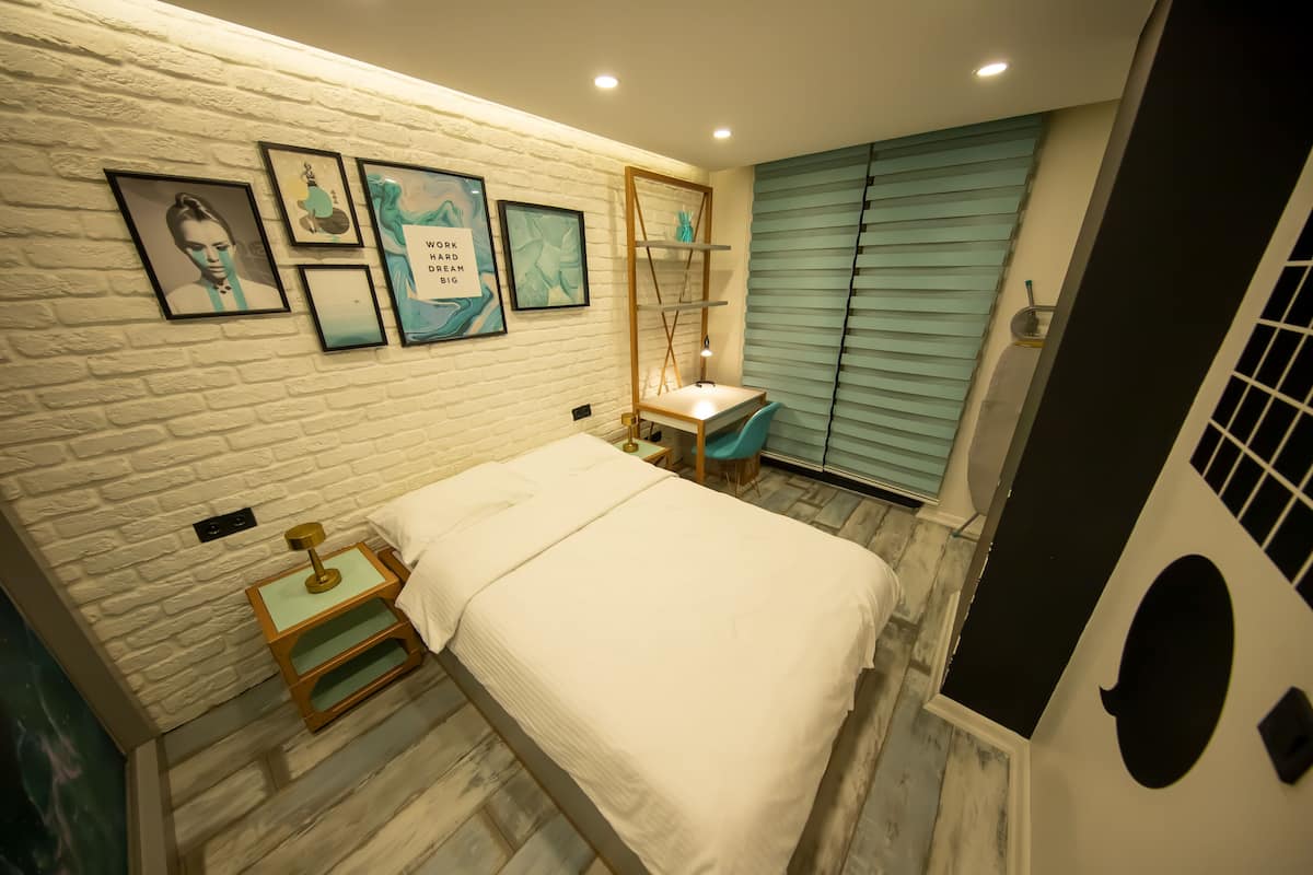 Un dormitorio con pared de ladrillo blanco, cama con ropa de cama blanca, mesita de noche con lámpara y escritorio con silla.