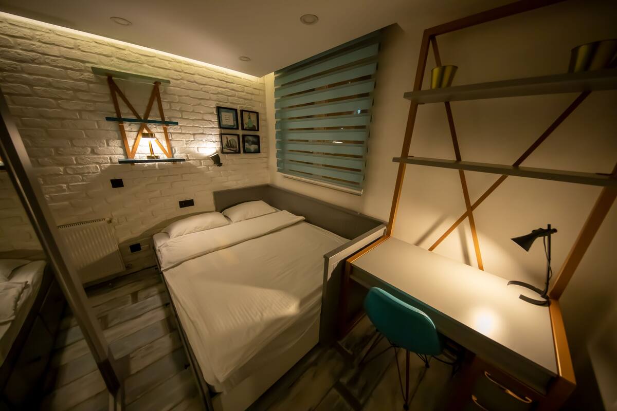 Un dormitorio compacto con una cama individual sobre otra, un escritorio con silla, una estantería y una ventana con persianas.