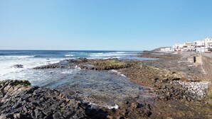 A rocky shoreline with waves, a clear sky, and buildings in the background.