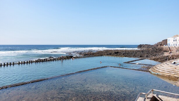 A coastal swimming area with clear water, rocky shore, and a building with steps leading down to the water.