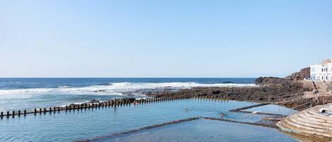 A coastal swimming area with clear water, rocky shore, and a building with steps leading down to the water.