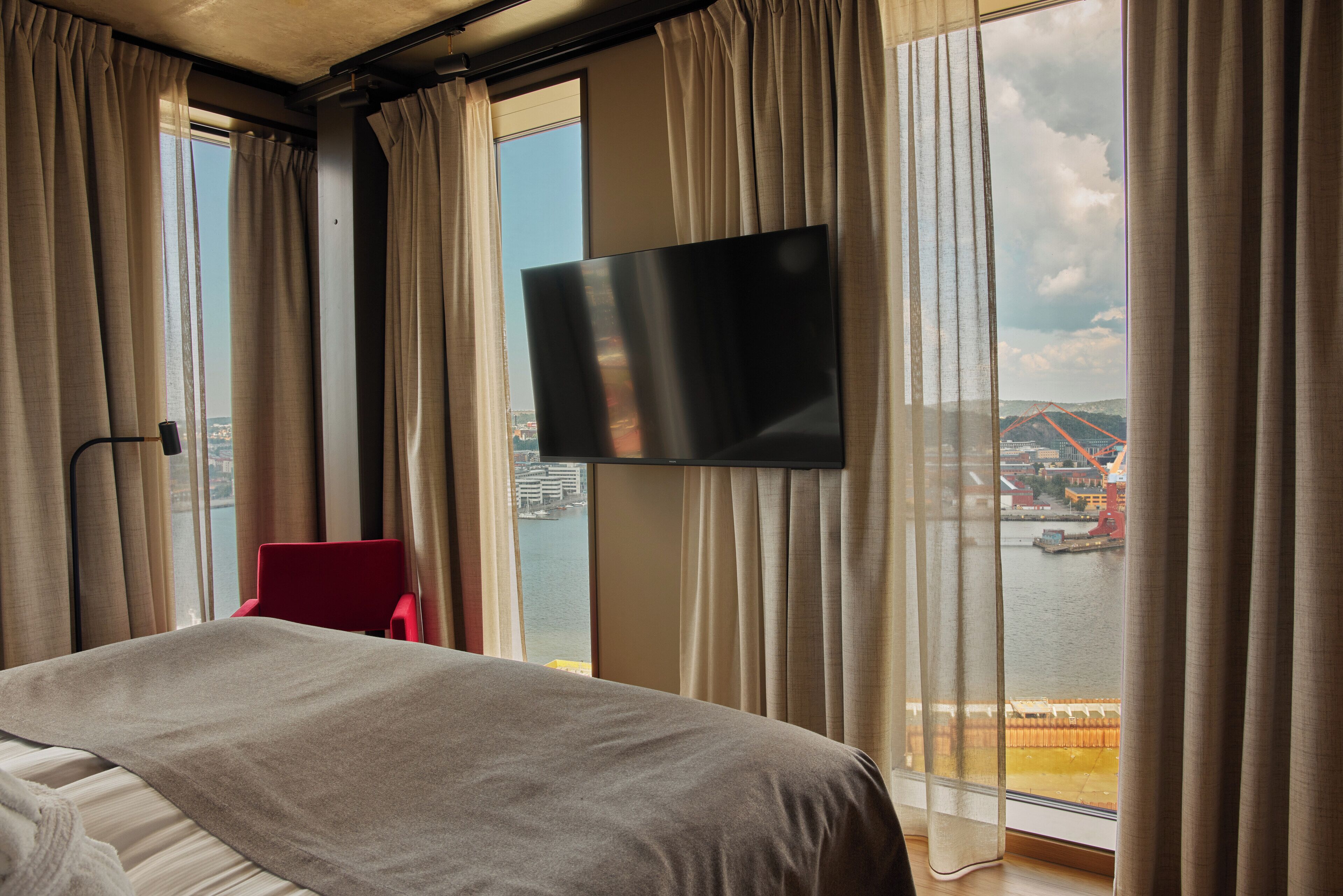 A hotel room with a bed, a red chair, a TV mounted on the wall, and a view of a bridge and water.