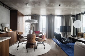 Een moderne hotelkamer met een ronde eettafel, meerdere stoelen, een bank, een televisie en een groot raam met gordijnen.