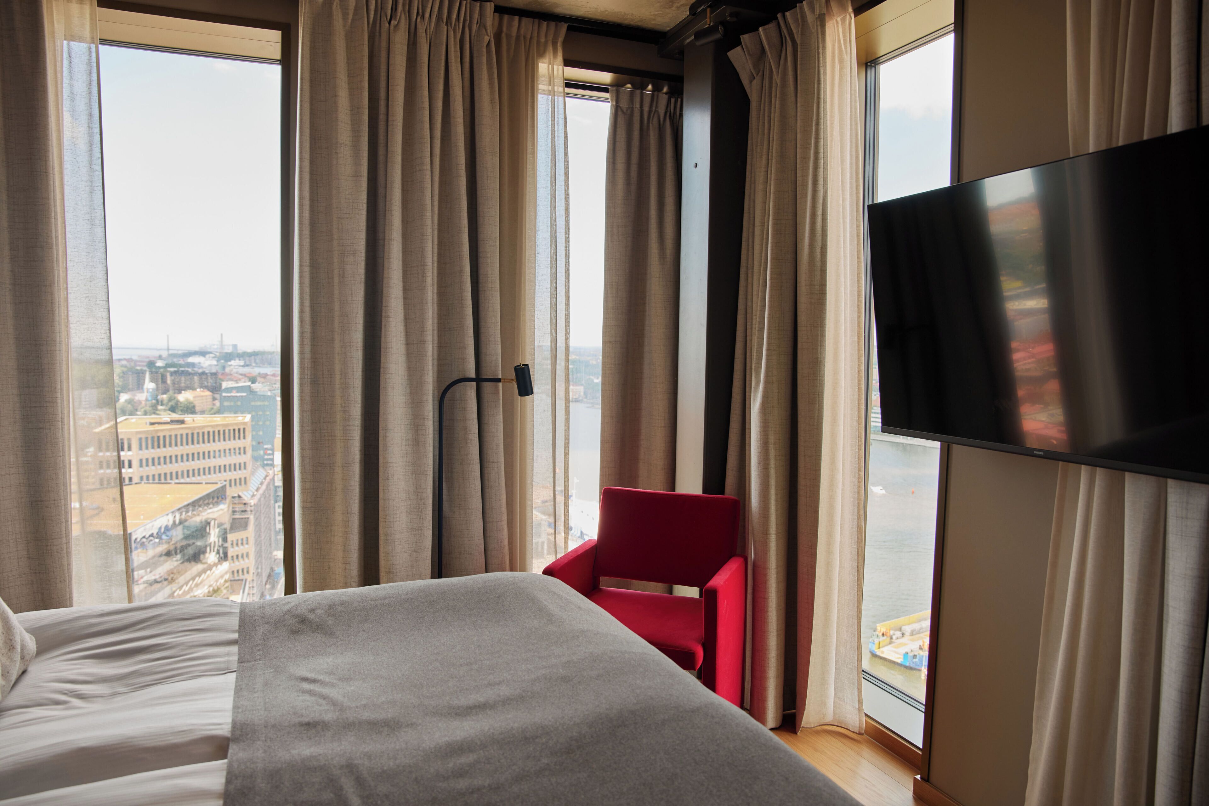 Una habitación de hotel con una cama, una silla roja, un televisor y un ventanal con cortinas.