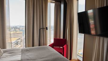 Una habitación de hotel con una cama, una silla roja, un televisor y un ventanal con cortinas.