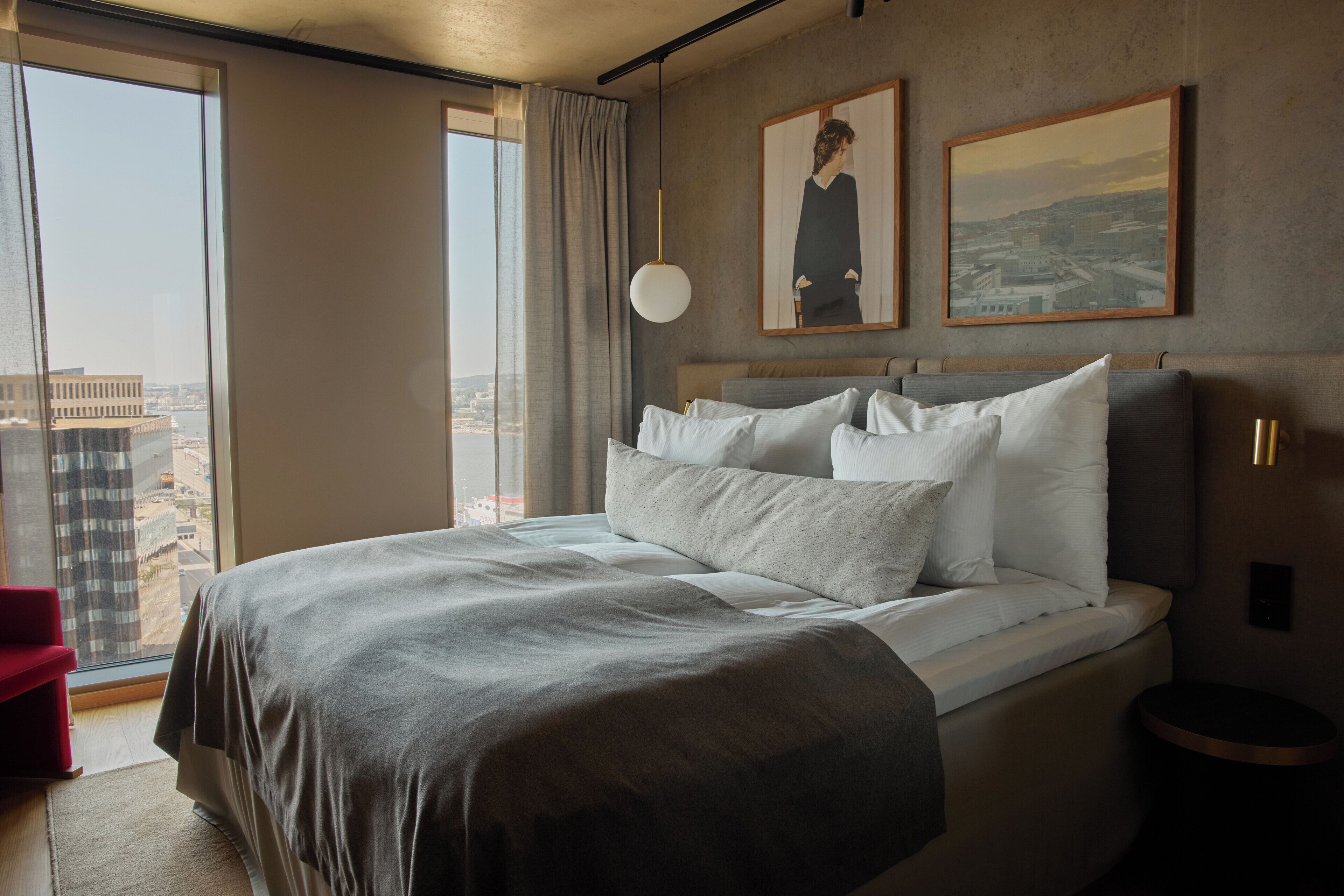 A modern hotel room with a large bed, a view of the city, and two framed pictures on the wall.