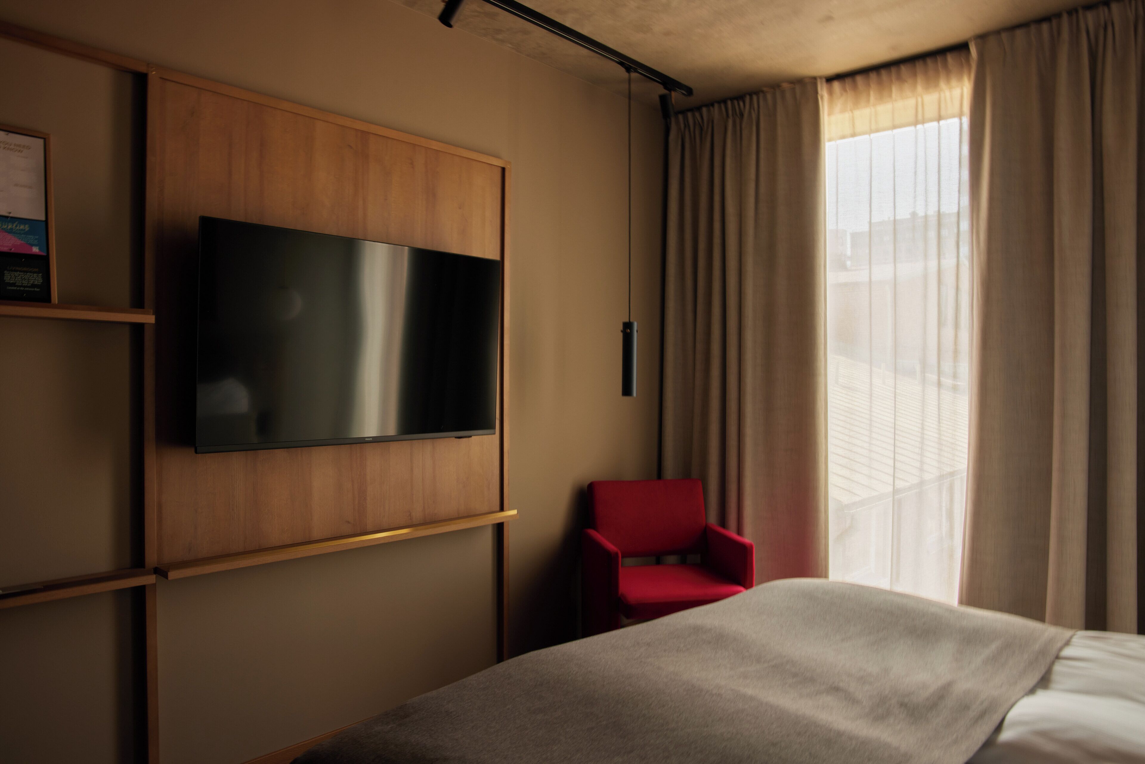 A hotel room with a bed, a red chair, a flat-screen TV mounted on a wooden panel, and a large window with curtains.