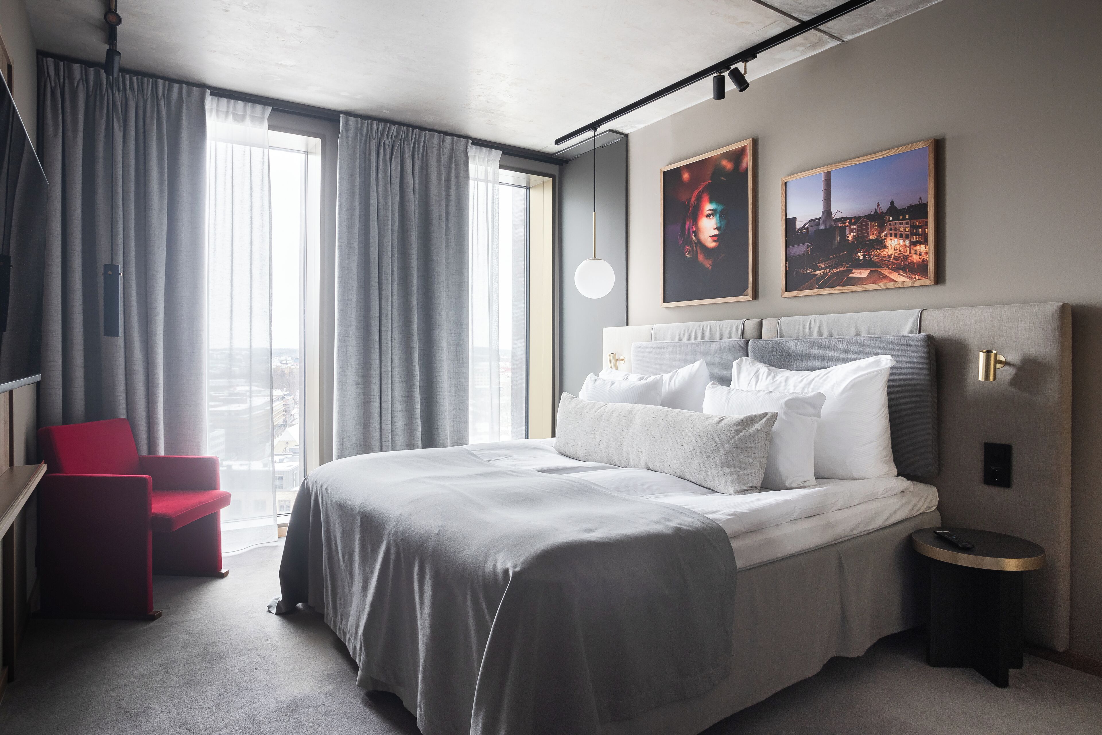 Une chambre d’hôtel moderne avec un grand lit, un fauteuil rouge, une petite table ronde et deux tableaux encadrés accrochés au mur.
