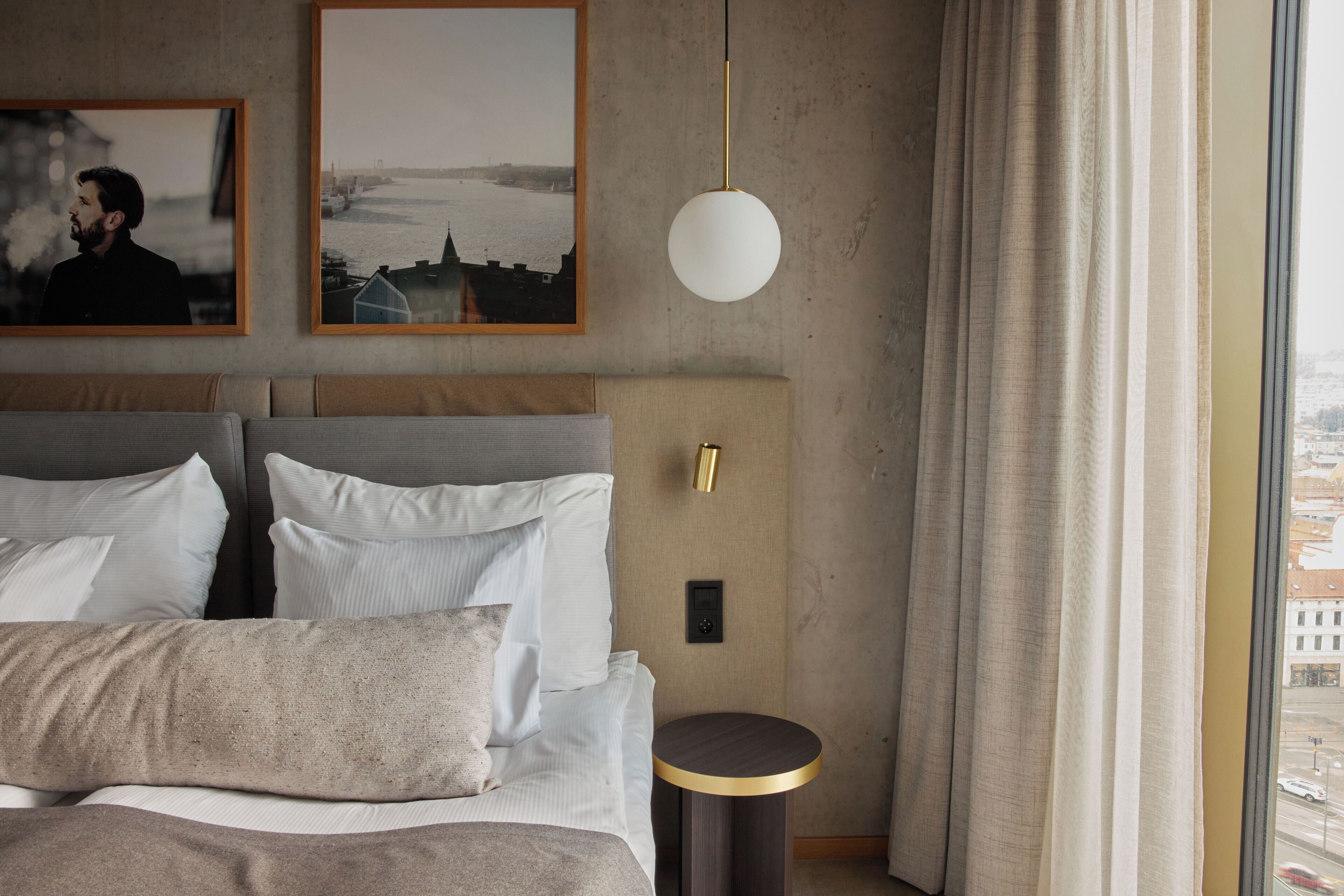 A modern hotel room with a large bed, a round side table, a wall-mounted light, and two framed pictures.