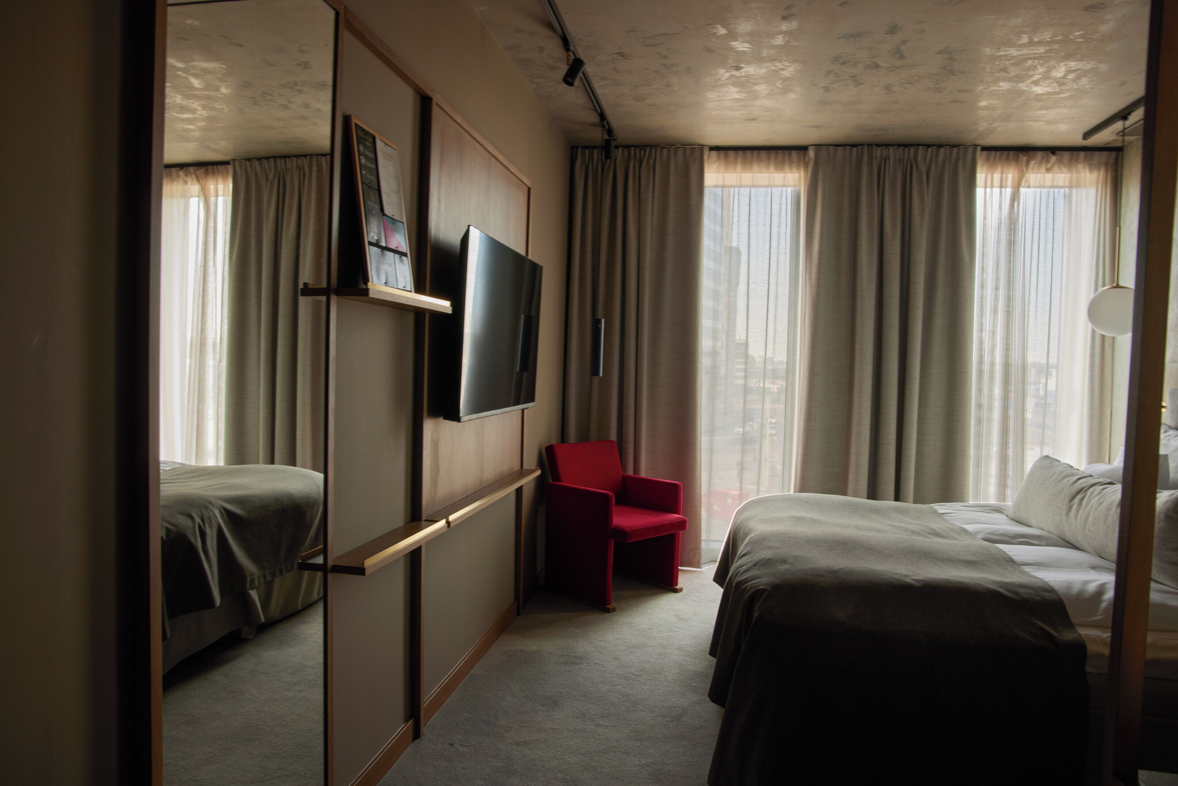 Une chambre d’hôtel avec deux lits, un téléviseur fixé au mur, un fauteuil rouge et de grandes fenêtres avec des rideaux.