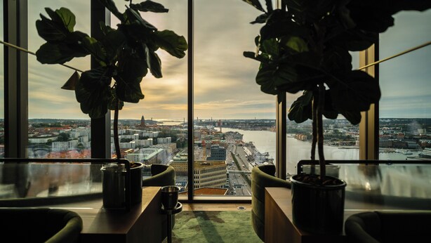 Habitación con ventanas amplias que ofrecen vista a la ciudad, dos plantas en macetas y una pequeña mesa con una taza.