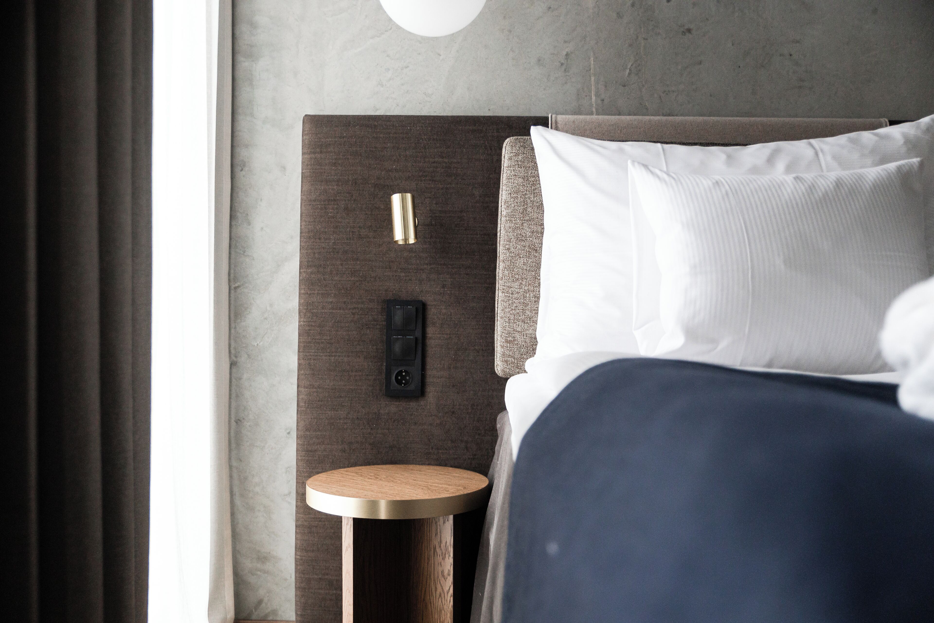 Une chambre d’hôtel moderne, équipée d’un lit, d’une table de chevet ronde en bois et d’un panneau de commande fixé au mur.